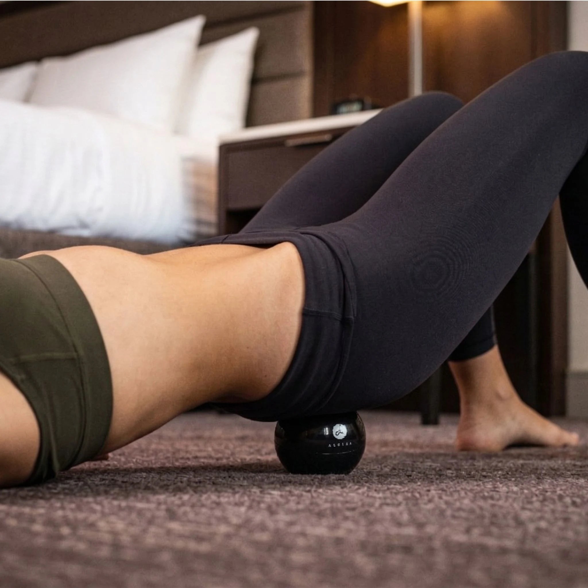Person using a black massage Orbit ball on their glute in a bedroom setting